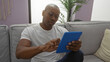 © Krakenimages.com - Young man sitting on couch in living room using tablet indoors at home wearing glasses and casual clothing, african american, relaxed in modern apartment setting.