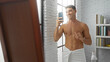 © Krakenimages.com - Young hispanic man smiling taking a selfie in front of a mirror wrapped in a towel in a modern living room with books and natural light streaming through a window.