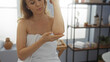 © Krakenimages.com - Woman applying skincare on elbow wrapped in towel in tranquil spa room with soft lighting and treatment products around, embodying wellness and beauty relaxation ambiance.