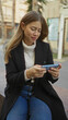 © Krakenimages.com - Young woman sitting outdoors in an urban street engaged with her smartphone wearing a coat and jeans looking focused with a slight smile, showcasing a casual city lifestyle.