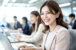 © Tondone - young woman in business suit smiles while working on laptop in modern office. Colleagues are engaged in their tasks, creating positive work environment