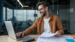 © dubogray1985 - The photo shows a smiling man with a beard and glasses sitting at a wooden desk in a modern office. He is wearing a brown jacket over a white shirt and is working on a laptop