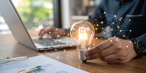 Poster - A person holds a glowing light bulb while using a laptop, symbolizing innovation and creativity in a modern workspace.