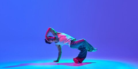 Skilled young dancer performing breathtaking breakdance tricks with against gradient blue purple background in neon light. Concept of hip-hop culture, modern street style, creativity, and movement.