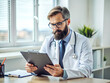 © Alisa - Concentrated doctor with beard and glasses is sitting at a desk and carefully reviewing patient information on a clipboard in his office