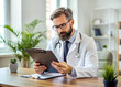 © Alisa - Concentrated doctor with beard and glasses is sitting at a desk and carefully reviewing patient information on a clipboard in his office