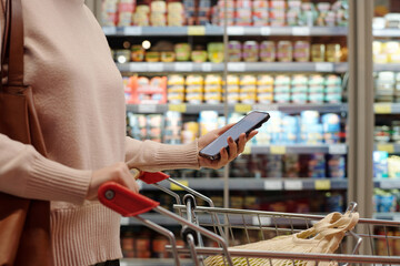 Side view of unrecognizable adult woman doing grocery shopping in supermarket and using phone while pushing cart, copy space