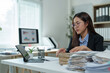 © crizzystudio - Young businesswoman organizing paperwork at her office desk, using a tablet and computer with charts displayed on the screen, efficiently managing her workload