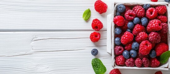 Poster - Close up of a white wooden background featuring a lunch box filled with vibrant raspberries and blueberries surrounded by mint leaves on the side