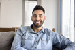 © fizkes - Portrait of happy Hispanic guy dressed in casual denim shirt looks at camera with joyful expression, spend day off alone seated on couch in cozy living room, participate in videocall, profile picture