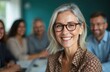© Vadym - Smiling mature businesswoman portrait in office with diverse colleagues in background. Senior woman looks at camera. Business meeting, teamwork, collaboration, leadership in modern company. Positive