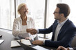 © fizkes - Successful partnership. Smiling young businessman in formal suit shake hand of happy senior female mentor experienced colleague sitting at boardroom table appreciate for help advice assistance in work