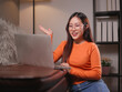 © Oil And Den - A woman wearing glasses in a orange sweater with laptop is sitting comfortably on sofa, video calling and  explaining with a smile.