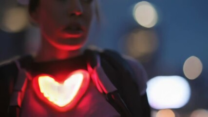 Poster - woman wearing futuristic heart shaped glowing device moving through dimly illuminated urban nightsca