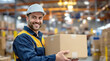 © vstudio - Smiling warehouse worker in safety gear holding cardboard box in distribution center