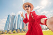 © EdNurg - Young woman wearing red dress and straw hat taking a selfie with skyscrapers in the background in Abu Dhabi