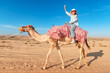 © EdNurg - Smiling female tourist riding a dromedary camel in the desert during a sunny day