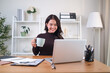 © wattana - A young businesswoman in a brown blazer and beige sweater is sitting at her home office desk, smiling as she works on her laptop and enjoys of coffee.
