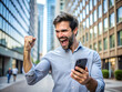 © Alisa - Man celebrating success while looking at smartphone. He is expressing happiness and excitement with fist pump. Urban background adds dynamic energy to his joyful reaction