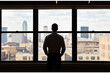 © Yulita - A businessman stands with hands in pockets looking out a large office window at a cityscape