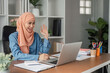 © wichayada - Cheerful Muslim woman in hijab waving during a video call in a modern office.