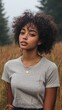 © indyntk - Portrait of a young woman with curly hair in a gray t-shirt standing in a serene field surrounded by tall grass and trees on a misty day