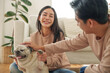 © eggeeggjiew - Asian couple sits on a comfortable floor, smiling and playing with their pet dog. joyful atmosphere together.