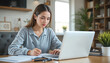 © aiartth - Young Caucasian woman doing homework and taking notes using a laptop at home, brainstorming ideas for her startup and working remotely.