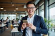 © SimpleDesignStudio - Smiling Young Asian Male Professional in Business Attire Using Smartphone in Modern Office Space with Colleagues Working in Background