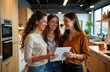 © Dipsky - Three women are gathered in a contemporary kitchen smiling and looking at a tablet. They appear engaged and excited possibly sharing recipes or cooking tips together.