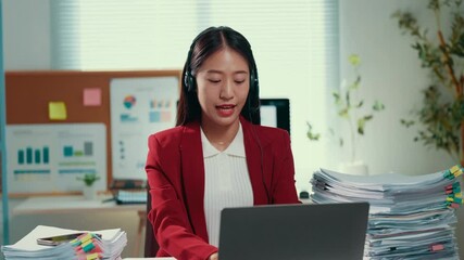 Poster - Young Asian businesswoman wearing red jacket and headset multitasking with laptop, efficiently managing paperwork in modern corporate workspace