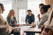 © Maskot - Smiling young entrepreneur discussing business strategy with male and female coworkers in meeting room at office
