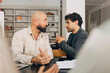 © Maskot - Young entrepreneur discussing with male colleague while sitting at table in meeting room at creative office