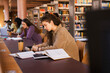 © Maskot - Young female student writing in book while sitting at desk studying inside university library