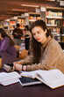 © Maskot - Young confident female university student writing in book while sitting at library desk