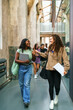 © Maskot - Full length of happy young female friends talking while walking in college corridor