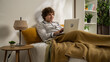 © kinomaster - Young man in the morning in bedroom sitting in bed in pajama, working on laptop, typing on keyboard, freelance work or surfing in the web.