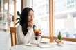 An asian woman is drinking the ice tea while looking through large window walls of the cafe.