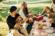 © polinaloves - Happy people having picnic with hot dogs and cold drinks, sitting on a plaid in a park.