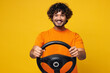 © ViDi Studio - Young smiling happy cheerful fun Indian man he wear orange t-shirt casual clothes hold steering wheel driving car looking camera isolated on plain yellow background studio portrait. Lifestyle concept.
