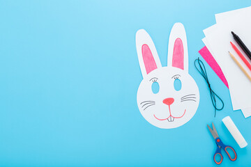 Wall Mural - White smiling rabbit paper mask on light blue table background. Pastel color. Closeup. Creating easter decoration for kid. Empty place for text. Top down view.