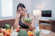 © Tj - Young sportswoman suffering headache while holding a bowl of salad and having healthy food such as vegetables and fruits on a table at home, healthcare and healthy food concept