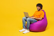 © ViDi Studio - Full body young smart IT Indian man he wear orange t-shirt casual clothes sit in bag chair hold use work on laptop pc computer isolated on plain yellow background studio portrait. Lifestyle concept