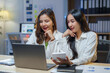 © amnaj - Professional Asian women analyzing financial data, leaning over laptop and paperwork in contemporary corporate workspace during evening hours