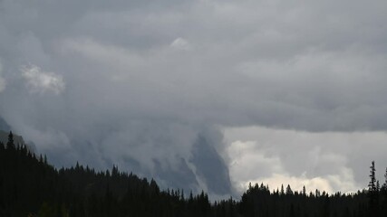 Wall Mural - Time lapse of gray storm rain clouds above a forest next with rocky mountain in the background
