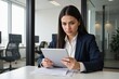 © MT - A focused young Hispanic woman in a business suit reviewing documents on a tablet in a modern office workspace, surrounded by computer desks and greenery.