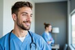 © MT - Cheerful Young Male Nurse in Blue Scrubs Wearing Stethoscope in Hospital Room with Female Colleague in Background, Focused on Patient Care and Compassionate Service