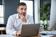 © Nina - Focused Young Caucasian Male Professional Working on Laptop in Modern Office Environment Surrounded by Indoor Plants and Papers