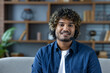 © Liubomir - Smiling man with curly hair wearing a headset in a modern living room during a video conference, looking directly at the camera.
