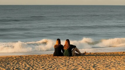 Poster - Female friends relaxing together on golden hued sandy beach, basking in warm sunlight while watching peaceful ocean waves during serene coastal sunset moment